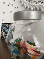 A clear glass jar with a metallic lid contains colorful gummy candies in various shapes, including a set resembling teeth. The jar is placed on a wooden surface. In the background, a decorative white frame is partially visible along with small googly eyes attached to the wall.