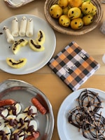 A collection of Halloween-themed snacks is displayed on a table. Bananas decorated as ghosts and pineapple slices drizzled with chocolate are on a white plate. Next to it, a basket contains oranges with jack-o'-lantern faces drawn on them. A checked napkin with black, orange, and white patterns lies nearby. Another dish has deviled eggs garnished with olives and sausages shaped like fingers. On another plate, pretzel and chocolate spider webs are artfully arranged.