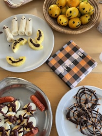 A collection of Halloween-themed snacks is displayed on a table. Bananas decorated as ghosts and pineapple slices drizzled with chocolate are on a white plate. Next to it, a basket contains oranges with jack-o'-lantern faces drawn on them. A checked napkin with black, orange, and white patterns lies nearby. Another dish has deviled eggs garnished with olives and sausages shaped like fingers. On another plate, pretzel and chocolate spider webs are artfully arranged.