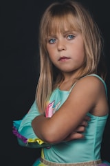 A young girl with long blonde hair and blue eyes stands with her arms crossed, wearing a colorful outfit in shades of teal, yellow, and pink. The background is dark, emphasizing her serious expression.