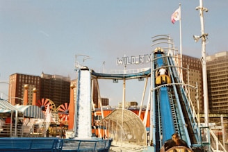 A vibrant amusement park scene featuring a water ride called Wild River. The structure is prominently blue with people in yellow boats ascending a ramp. Surrounding the ride are colorful tents and attractions. Tall buildings and a clear sky are visible in the background.