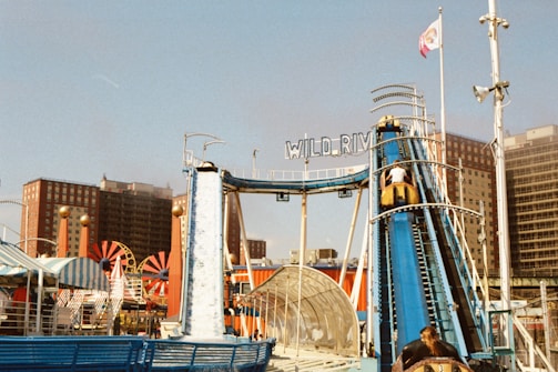 A vibrant amusement park scene featuring a water ride called Wild River. The structure is prominently blue with people in yellow boats ascending a ramp. Surrounding the ride are colorful tents and attractions. Tall buildings and a clear sky are visible in the background.