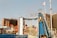 A vibrant amusement park scene featuring a water ride called Wild River. The structure is prominently blue with people in yellow boats ascending a ramp. Surrounding the ride are colorful tents and attractions. Tall buildings and a clear sky are visible in the background.