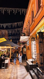 A lively street scene at night with two-story buildings adorned with decorative lights. People are walking along the cobblestone path, and some are seated under a canopy at a bar with a cozy ambiance. The warm glow from the streetlights and store signs adds a welcoming atmosphere.