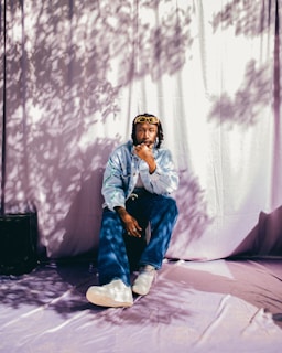 A person sits thoughtfully on a purple cloth backdrop, wearing a light denim jacket, blue jeans, and white sneakers. The scene is highlighted by dappled sunlight and shadow patterns created by tree leaves.