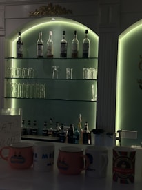 A dimly lit bar with illuminated shelves containing multiple bottles of alcohol and glassware. The bottom shelf displays various liquor bottles, while several mugs with different designs are placed on the counter.