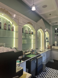 A well-lit bar area featuring a classic design with illuminated shelves displaying an assortment of liquor bottles. The shelves are framed by white columns and gold ornamental details. The countertop includes a coffee machine and bar equipment, with a patterned black and white floor. Hanging pendant lights and a high ceiling contribute to the sophisticated ambience.