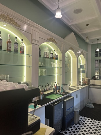 A well-lit bar area featuring a classic design with illuminated shelves displaying an assortment of liquor bottles. The shelves are framed by white columns and gold ornamental details. The countertop includes a coffee machine and bar equipment, with a patterned black and white floor. Hanging pendant lights and a high ceiling contribute to the sophisticated ambience.