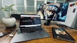 A workspace with a wooden desk featuring a laptop and an external monitor displaying a digital image of a person wearing a traditional headdress. There is also a potted plant and a closed book titled 'Unposted Letters' on the desk. Cables and a mouse are visible near the laptop, and there's a whiteboard in the background.