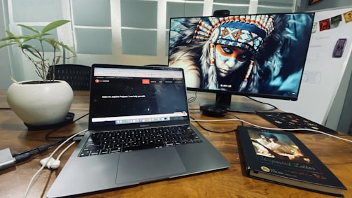 A workspace with a wooden desk featuring a laptop and an external monitor displaying a digital image of a person wearing a traditional headdress. There is also a potted plant and a closed book titled 'Unposted Letters' on the desk. Cables and a mouse are visible near the laptop, and there's a whiteboard in the background.