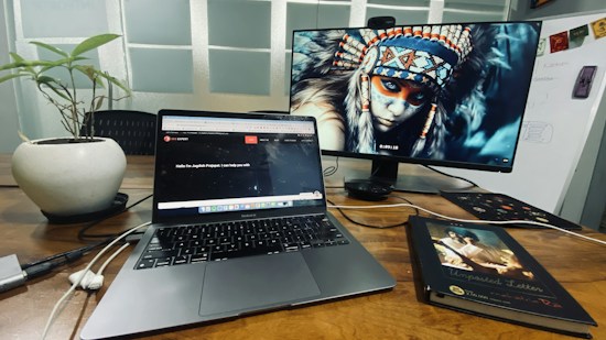 A workspace with a wooden desk featuring a laptop and an external monitor displaying a digital image of a person wearing a traditional headdress. There is also a potted plant and a closed book titled 'Unposted Letters' on the desk. Cables and a mouse are visible near the laptop, and there's a whiteboard in the background.
