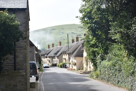 A picturesque village street lined with charming thatched-roof cottages, bordered by lush greenery. The road gently curves with trees and hedges along one side. In the background, a hill provides a scenic backdrop under a clear sky.