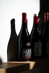 A row of wine bottles stands on a wooden shelf, each bottle casting distinct shadows on the white wall behind. One bottle is prominently labeled 'Saint Louis' with a red cap.