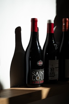 A row of wine bottles stands on a wooden shelf, each bottle casting distinct shadows on the white wall behind. One bottle is prominently labeled 'Saint Louis' with a red cap.
