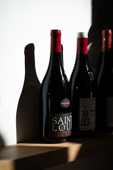 A row of wine bottles stands on a wooden shelf, each bottle casting distinct shadows on the white wall behind. One bottle is prominently labeled 'Saint Louis' with a red cap.