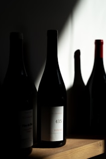 Several wine bottles are placed on a wooden shelf, with one prominently displaying a label indicating '43.5 Syrah'. The lighting creates dramatic shadows, highlighting the textures and shapes of the bottles.