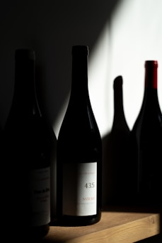 Several wine bottles are placed on a wooden shelf, with one prominently displaying a label indicating '43.5 Syrah'. The lighting creates dramatic shadows, highlighting the textures and shapes of the bottles.