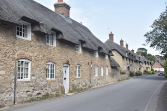 A row of traditional stone cottages with thatched roofs lines a narrow, quiet street. The cottages feature white-framed windows and chimneys. Lush greenery and trees surround the area, and a red car is parked further down the road.