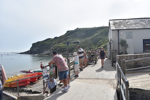 People are gathered near a coastal area with a rocky hill and calm sea. Several kayaks are lined up on the beach, and a building marked 'Boat Shed' is visible. The weather appears clear and sunny, with some standing and leaning against barriers.