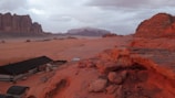 An expansive desert landscape featuring rugged red rock formations and sandy plains under a cloudy sky. In the foreground, there are several structures resembling tents or cabins, built with dark roofs and striped pattern walls. The scene captures the vastness and natural beauty of the desert environment with distant mountains visible on the horizon.