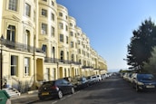 A row of elegant, yellow townhouses with ornate black railings line a street filled with parked cars on either side. The sky is clear and blue, suggesting a bright, sunny day. A few trees are visible on the right, adding greenery to the urban setting.