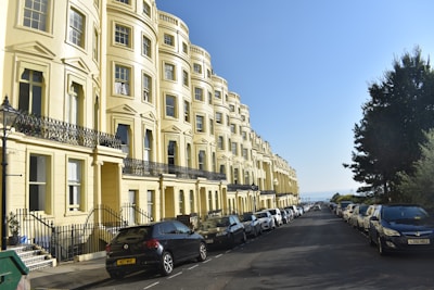 A row of elegant, yellow townhouses with ornate black railings line a street filled with parked cars on either side. The sky is clear and blue, suggesting a bright, sunny day. A few trees are visible on the right, adding greenery to the urban setting.