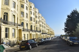 A row of elegant, yellow townhouses with ornate black railings line a street filled with parked cars on either side. The sky is clear and blue, suggesting a bright, sunny day. A few trees are visible on the right, adding greenery to the urban setting.