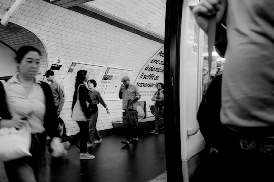 People are walking and standing in a subway station, with some examining a map or advertisement on the tiled wall. The atmosphere is bustling and busy, typical of urban transit environments.