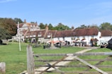 A picturesque countryside setting features a charming rustic building with a brick roof and white walls, adorned with hanging flower baskets. It is surrounded by a well-maintained grassy area. Outdoor seating with people enjoying the day can be seen under colorful umbrellas. A wooden gate and a flagpole with a Union Jack flag complete the scene.