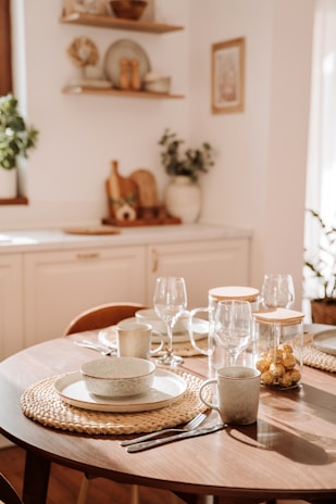A wooden dining table is elegantly set with ceramic dishes, cups, and transparent wine glasses on woven placemats. A jar of golden-wrapped chocolates adds a touch of luxury. The kitchen area features white cabinetry and decorative shelves with plants and kitchenware, illuminated by warm natural light.