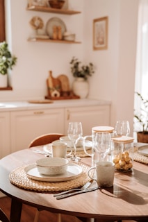 A wooden dining table is elegantly set with ceramic dishes, cups, and transparent wine glasses on woven placemats. A jar of golden-wrapped chocolates adds a touch of luxury. The kitchen area features white cabinetry and decorative shelves with plants and kitchenware, illuminated by warm natural light.