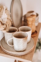 A set of rustic ceramic cups and plates are neatly arranged on a wooden shelf. The texture gives a handcrafted, artisanal feel. In the background, there are additional kitchen items, including a pepper grinder and a stack of bowls, adding to the cozy, homey atmosphere.
