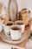 A set of rustic ceramic cups and plates are neatly arranged on a wooden shelf. The texture gives a handcrafted, artisanal feel. In the background, there are additional kitchen items, including a pepper grinder and a stack of bowls, adding to the cozy, homey atmosphere.