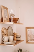 Two wooden shelves are mounted on a wall, displaying various home decor items. The top shelf holds a framed photo, a wine glass, a wooden bowl, and a tall candle. The lower shelf features stacked ceramic plates and bowls, a mug, a wooden pepper grinder, a sponge, and a soap bar.