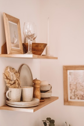 Two wooden shelves are mounted on a wall, displaying various home decor items. The top shelf holds a framed photo, a wine glass, a wooden bowl, and a tall candle. The lower shelf features stacked ceramic plates and bowls, a mug, a wooden pepper grinder, a sponge, and a soap bar.