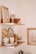 Two wooden shelves are mounted on a wall, displaying various home decor items. The top shelf holds a framed photo, a wine glass, a wooden bowl, and a tall candle. The lower shelf features stacked ceramic plates and bowls, a mug, a wooden pepper grinder, a sponge, and a soap bar.