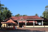 A traditional Indian temple with a terracotta-tiled roof and supporting columns, surrounded by lush greenery and palm trees. Several people are seen standing near the entrance, some wearing traditional attire.