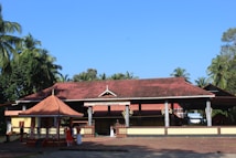 A traditional Indian temple with a terracotta-tiled roof and supporting columns, surrounded by lush greenery and palm trees. Several people are seen standing near the entrance, some wearing traditional attire.