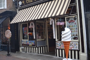 A storefront with a striped awning characterized by its candy-themed decorations. Large candy canes flank the entrance, and a giant ice cream cone stands beside a window displaying various colorful posters. Inside, shelves filled with candy are visible.