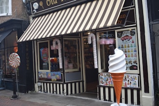 A storefront with a striped awning characterized by its candy-themed decorations. Large candy canes flank the entrance, and a giant ice cream cone stands beside a window displaying various colorful posters. Inside, shelves filled with candy are visible.
