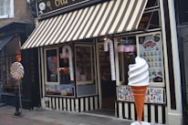 A storefront with a striped awning characterized by its candy-themed decorations. Large candy canes flank the entrance, and a giant ice cream cone stands beside a window displaying various colorful posters. Inside, shelves filled with candy are visible.