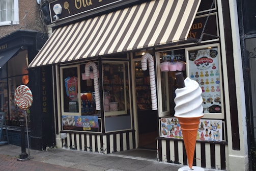 A storefront with a striped awning characterized by its candy-themed decorations. Large candy canes flank the entrance, and a giant ice cream cone stands beside a window displaying various colorful posters. Inside, shelves filled with candy are visible.