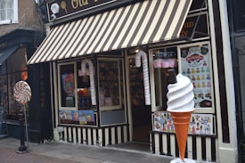 A storefront with a striped awning characterized by its candy-themed decorations. Large candy canes flank the entrance, and a giant ice cream cone stands beside a window displaying various colorful posters. Inside, shelves filled with candy are visible.