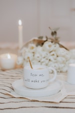 A cozy scene featuring a white coffee mug with the inscription 'Dear me, I will make you proud' placed on a saucer. Background elements include a bouquet of white flowers, lit candles, and an open book beneath the saucer. Everything is laid out on a striped fabric.