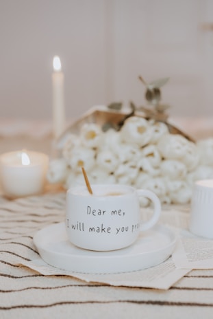 A cozy scene featuring a white coffee mug with the inscription 'Dear me, I will make you proud' placed on a saucer. Background elements include a bouquet of white flowers, lit candles, and an open book beneath the saucer. Everything is laid out on a striped fabric.