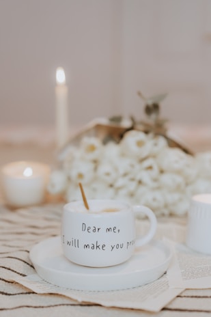 A white mug with an inspirational quote, 'Dear me, I will make you proud,' sits on a matching saucer. The setting includes a cozy arrangement with lit candles and a soft floral bouquet in the background, giving a warm, serene ambiance.