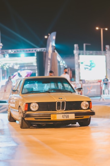 A classic BMW car with a Dubai license plate is parked in an outdoor venue under artificial lighting. The background includes people and event structures, with bright lights and banners partially visible.