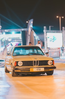 A classic BMW car with a Dubai license plate is parked in an outdoor venue under artificial lighting. The background includes people and event structures, with bright lights and banners partially visible.
