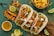 Three tacos filled with crispy fried pieces of fish, garnished with chopped vegetables, cream sauce, and served with lime wedges. A bowl of pineapple chunks and a small cup of dipping sauce are also present alongside grilled pineapple slices on a wooden board.