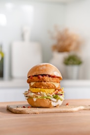 A gourmet burger stacked with various layers including fried components, cheese, a slice of pineapple, lettuce, and sauce. It sits on a wooden board in a softly lit kitchen setting, suggesting a carefully crafted and appetizing meal.